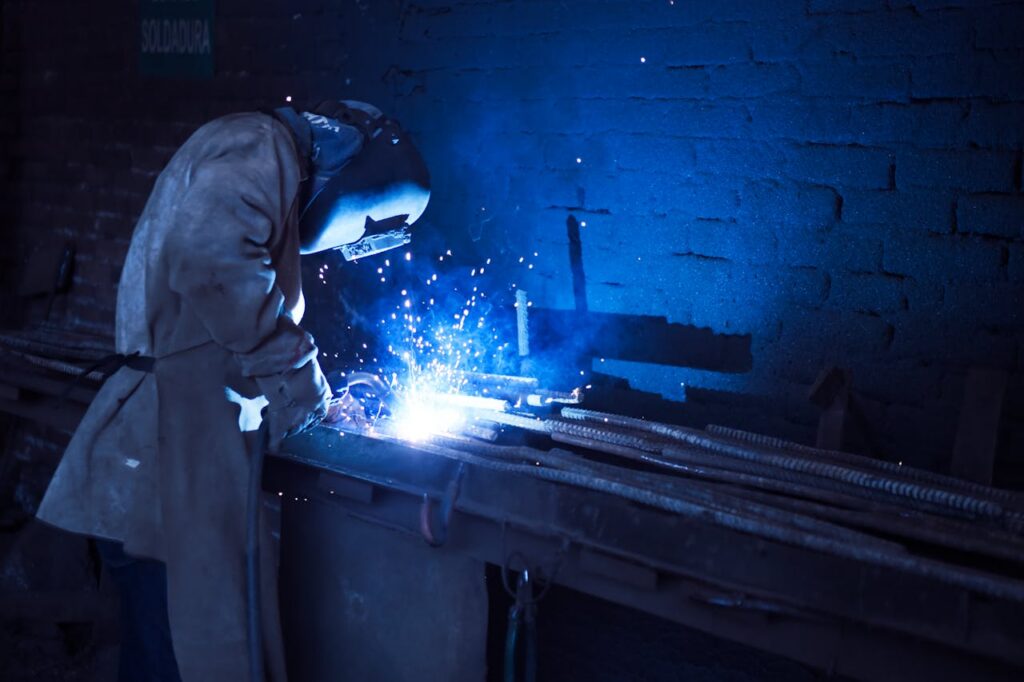 pexels-photo-1474993 Industrial worker welding metal indoors, emitting bright blue sparks and smoke.